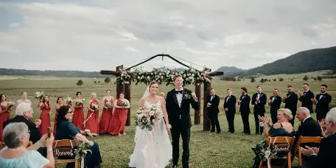 A bride and groom walking down the aisle at an outdoor wedding ceremony in a grassy field with mountains in the background.