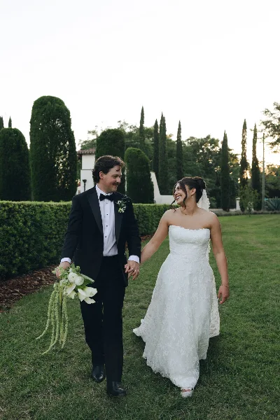 A bride and groom walking hand-in-hand on a grassy lawn with manicured hedges and trees in the background.