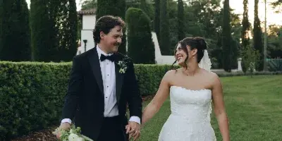 A bride and groom walking hand-in-hand on a grassy lawn with manicured hedges and trees in the background.