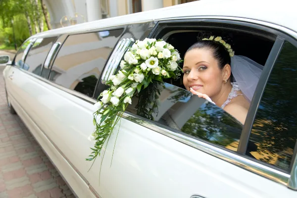 A bride in a white dress and veil sits inside a white stretch limousine decorated with a white floral arrangement.