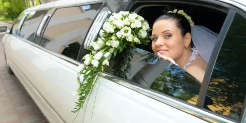 A bride in a white dress and veil sits inside a white stretch limousine decorated with a white floral arrangement.