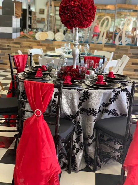 A decorated event table with black Chiavari chairs, red sashes, patterned tablecloth, and a red rose centerpiece.
