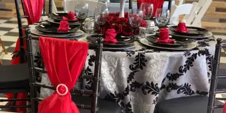 A decorated event table with black Chiavari chairs, red sashes, patterned tablecloth, and a red rose centerpiece.