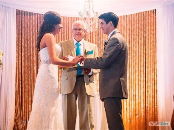 A wedding officiant stands between a bride and groom as they exchange rings during a ceremony.