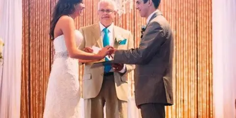 A wedding officiant stands between a bride and groom as they exchange rings during a ceremony.
