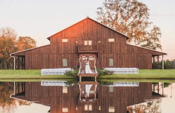 Exterior view of a large wooden barn wedding venue with a dock over a pond and white ceremony chairs set up on the lawn.
