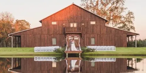Exterior view of a large wooden barn wedding venue with a dock over a pond and white ceremony chairs set up on the lawn.