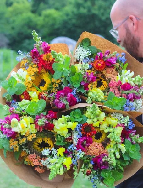 A collection of colorful wildflower bouquets wrapped in brown paper, featuring sunflowers, dahlias, and snapdragons.