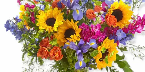 A colorful floral arrangement featuring sunflowers, irises, and roses in a clear glass vase.