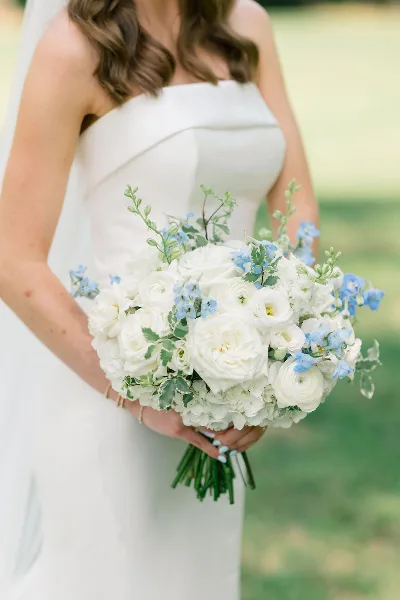 A bride in a white strapless gown holding a lush bouquet of white roses, ranunculus, and blue delphiniums.