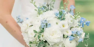 A bride in a white strapless gown holding a lush bouquet of white roses, ranunculus, and blue delphiniums.