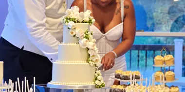 A bride and groom cutting a white tiered wedding cake decorated with white flowers at a wedding reception.