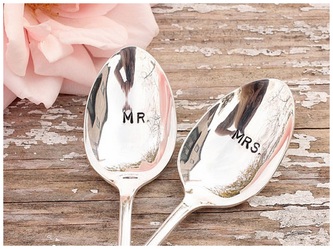 Two silver spoons engraved with MR and MRS beside a pale pink flower on a rustic wooden surface