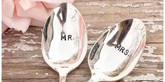 Two silver spoons engraved with MR and MRS beside a pale pink flower on a rustic wooden surface