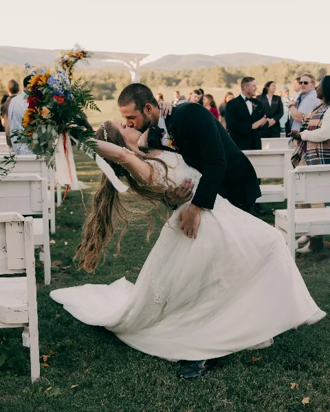 A groom dips his bride for a kiss during an outdoor wedding ceremony with guests watching in the background.