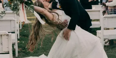 A groom dips his bride for a kiss during an outdoor wedding ceremony with guests watching in the background.