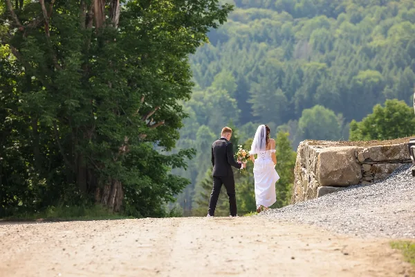 A bride and groom walking on a gravel path with a backdrop of lush green mountains and a stone wall.