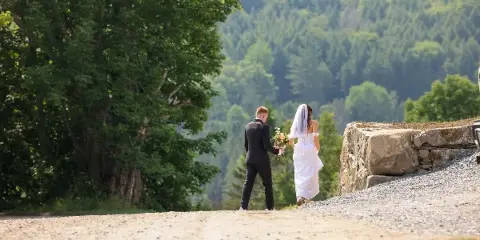 A bride and groom walking on a gravel path with a backdrop of lush green mountains and a stone wall.