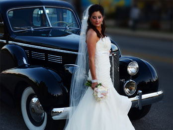 A bride in a white wedding gown and veil standing beside a vintage black car.