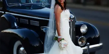 A bride in a white wedding gown and veil standing beside a vintage black car.