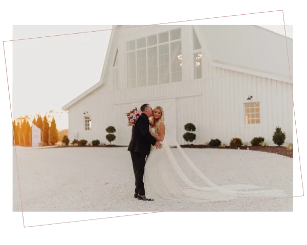 A bride and groom kissing in front of a large white barn wedding venue with a gravel courtyard.