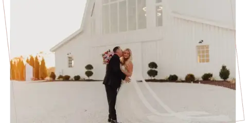 A bride and groom kissing in front of a large white barn wedding venue with a gravel courtyard.