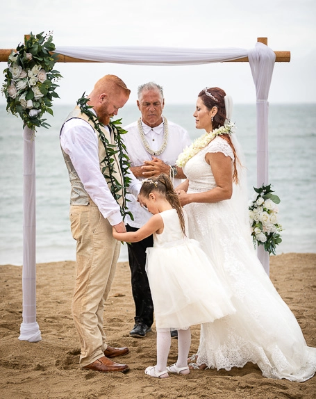 A wedding ceremony on a beach with a bride, groom, officiant, and flower girl under a floral arch.