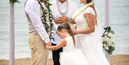 A wedding ceremony on a beach with a bride, groom, officiant, and flower girl under a floral arch.