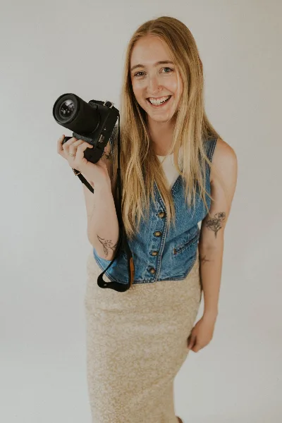 A smiling woman with long blonde hair holding a professional camera with a large lens.