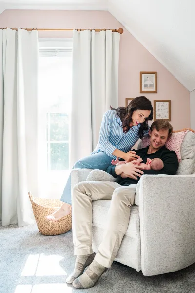 A mother and father smiling while holding their newborn baby in a living room chair.