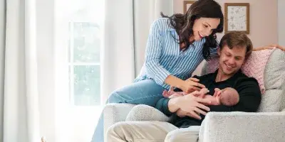 A mother and father smiling while holding their newborn baby in a living room chair.