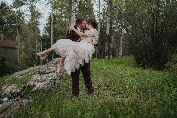 A groom in a maroon suit lifts and kisses his bride in a white dress in a grassy, wooded area.