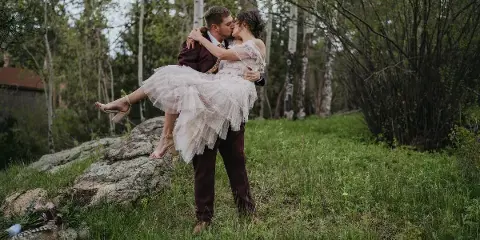A groom in a maroon suit lifts and kisses his bride in a white dress in a grassy, wooded area.