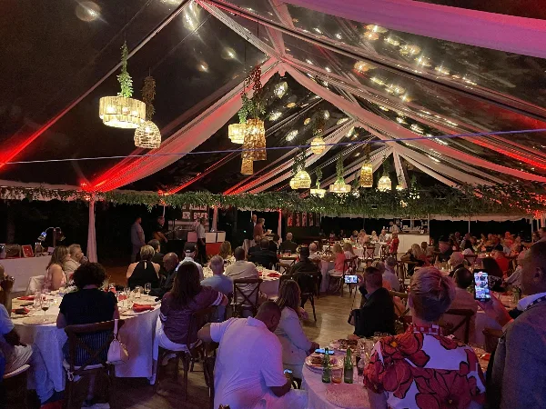 Interior of a large event tent with hanging pendant lights, greenery garlands, and guests seated at round tables.
