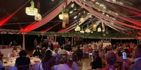 Interior of a large event tent with hanging pendant lights, greenery garlands, and guests seated at round tables.