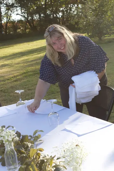 A woman setting a table with white linens and glassware outdoors.