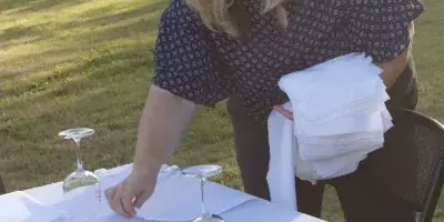 A woman setting a table with white linens and glassware outdoors.