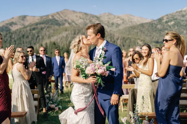 A bride and groom sharing a kiss during an outdoor wedding ceremony with guests clapping in the background.