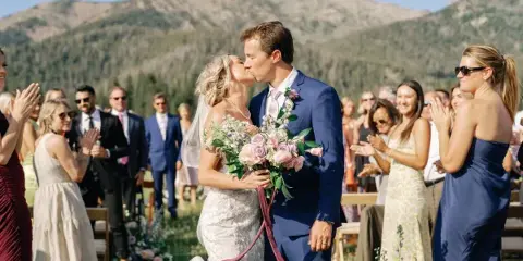 A bride and groom sharing a kiss during an outdoor wedding ceremony with guests clapping in the background.