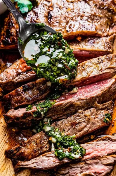 Sliced grilled meat with a fresh herb sauce being poured over it.
