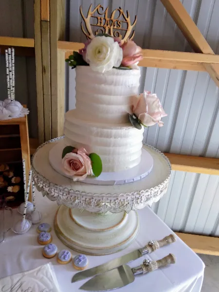 A three-tiered white wedding cake with buttercream texture, decorated with pink and white roses and a wooden topper, displayed on a cake stand.