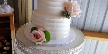 A three-tiered white wedding cake with buttercream texture, decorated with pink and white roses and a wooden topper, displayed on a cake stand.