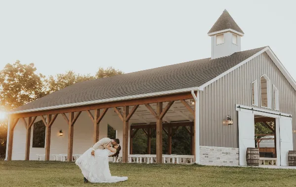 Exterior view of a large grey and white barn-style wedding venue with a covered porch and a couple in wedding attire in the foreground.