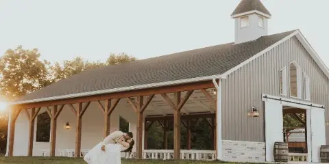 Exterior view of a large grey and white barn-style wedding venue with a covered porch and a couple in wedding attire in the foreground.