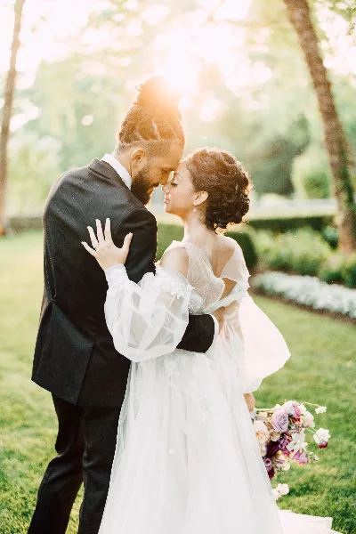 A bride and groom embracing outdoors in a garden setting with sunlight filtering through the trees.
