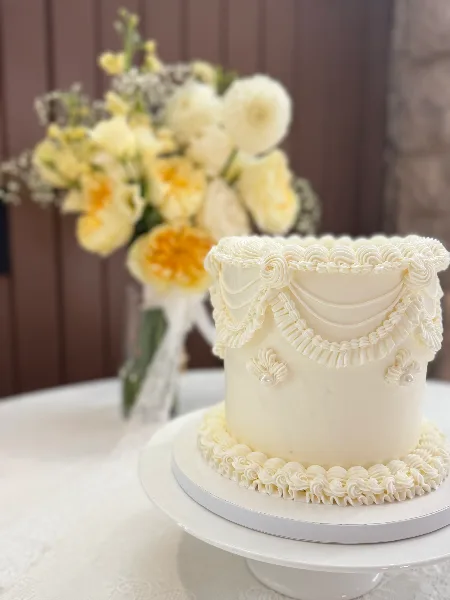 A white wedding cake with elaborate piped frosting details and pearl accents, displayed on a white cake stand next to a vase of yellow flowers.