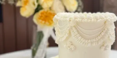 A white wedding cake with elaborate piped frosting details and pearl accents, displayed on a white cake stand next to a vase of yellow flowers.
