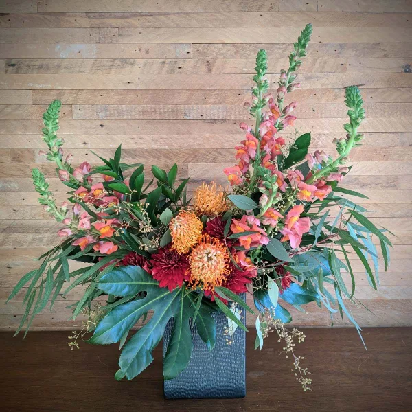 A colorful floral arrangement featuring orange pincushion proteas, red dahlias, and tall pink spikes in a textured blue vase against a wooden background.