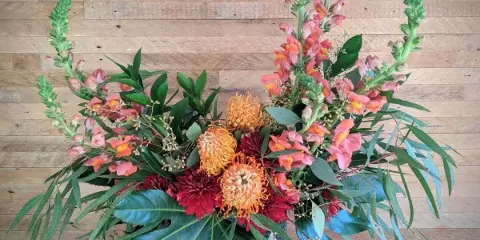 A colorful floral arrangement featuring orange pincushion proteas, red dahlias, and tall pink spikes in a textured blue vase against a wooden background.