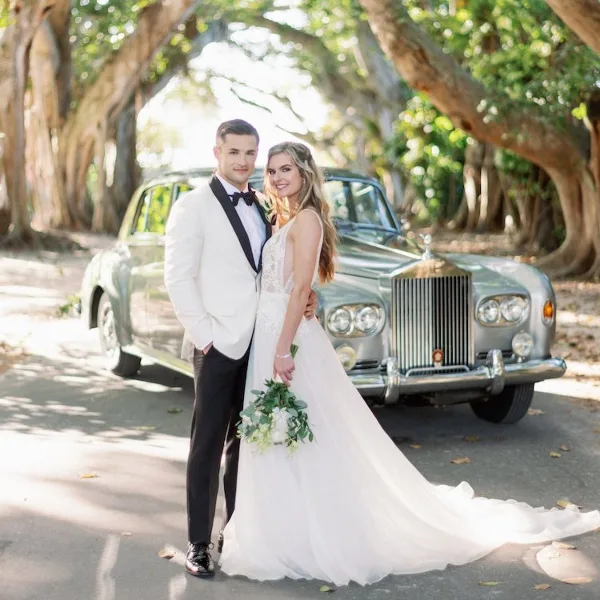 A bride and groom standing in front of a silver vintage Rolls-Royce car outdoors.